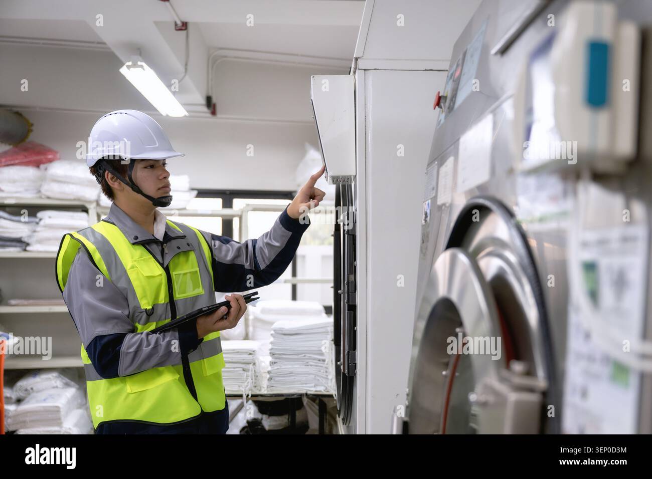 Il tecnico addetto alla manutenzione ispeziona le lavatrici di fabbrica indossano elmetto e gilet di sicurezza giallo fissano i punti della tavoletta alla macchina con pile di linee pulite Foto Stock