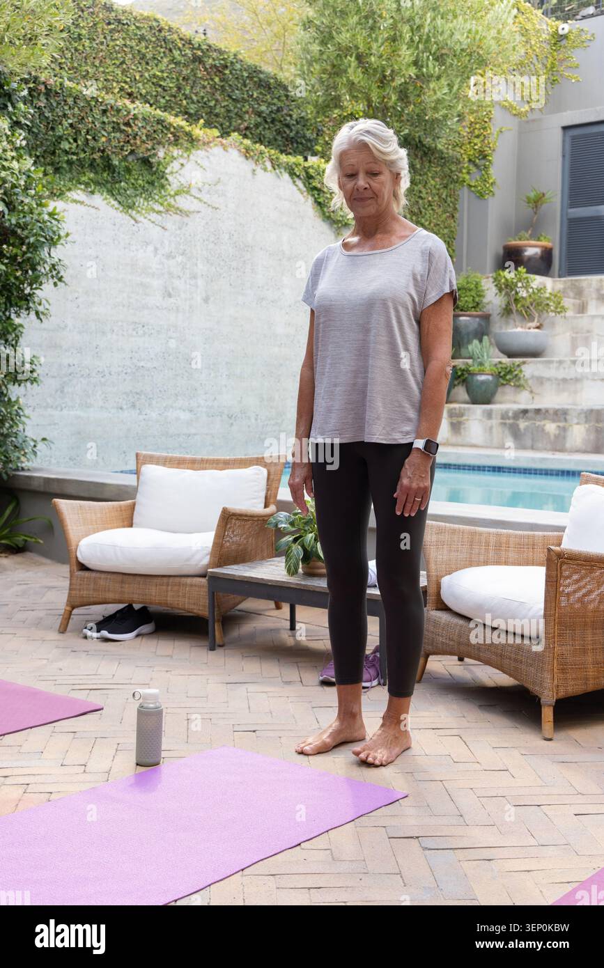 Donna anziana che indossa leggings e smartwatch, in piedi nudi sul patio a bordo piscina con tappetini viola Foto Stock