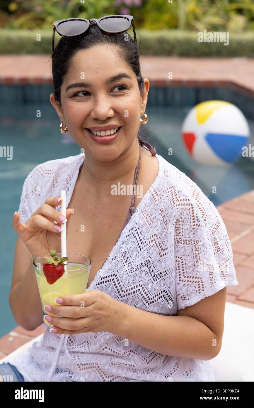 Donna seduta a bordo piscina che indossa una copertura in pizzo bianco, mescolando una bevanda gialla in vetro Foto Stock
