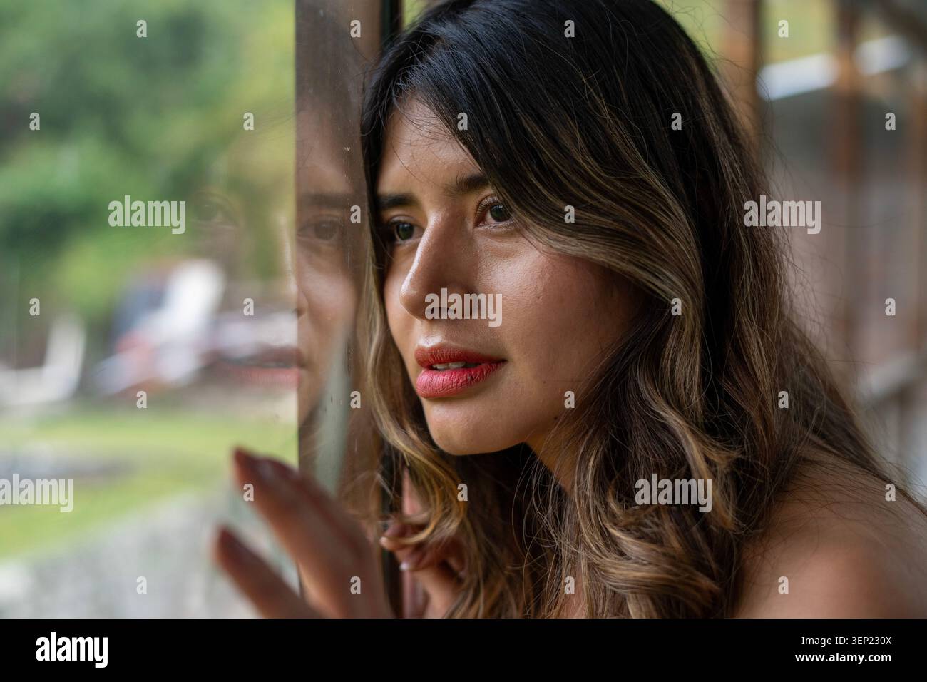 Giovane donna che guarda penosa, riflette sui pensieri e sulle emozioni, guarda attraverso una finestra di vetro con le mani riposate, mostrando un momento di disprezzo Foto Stock