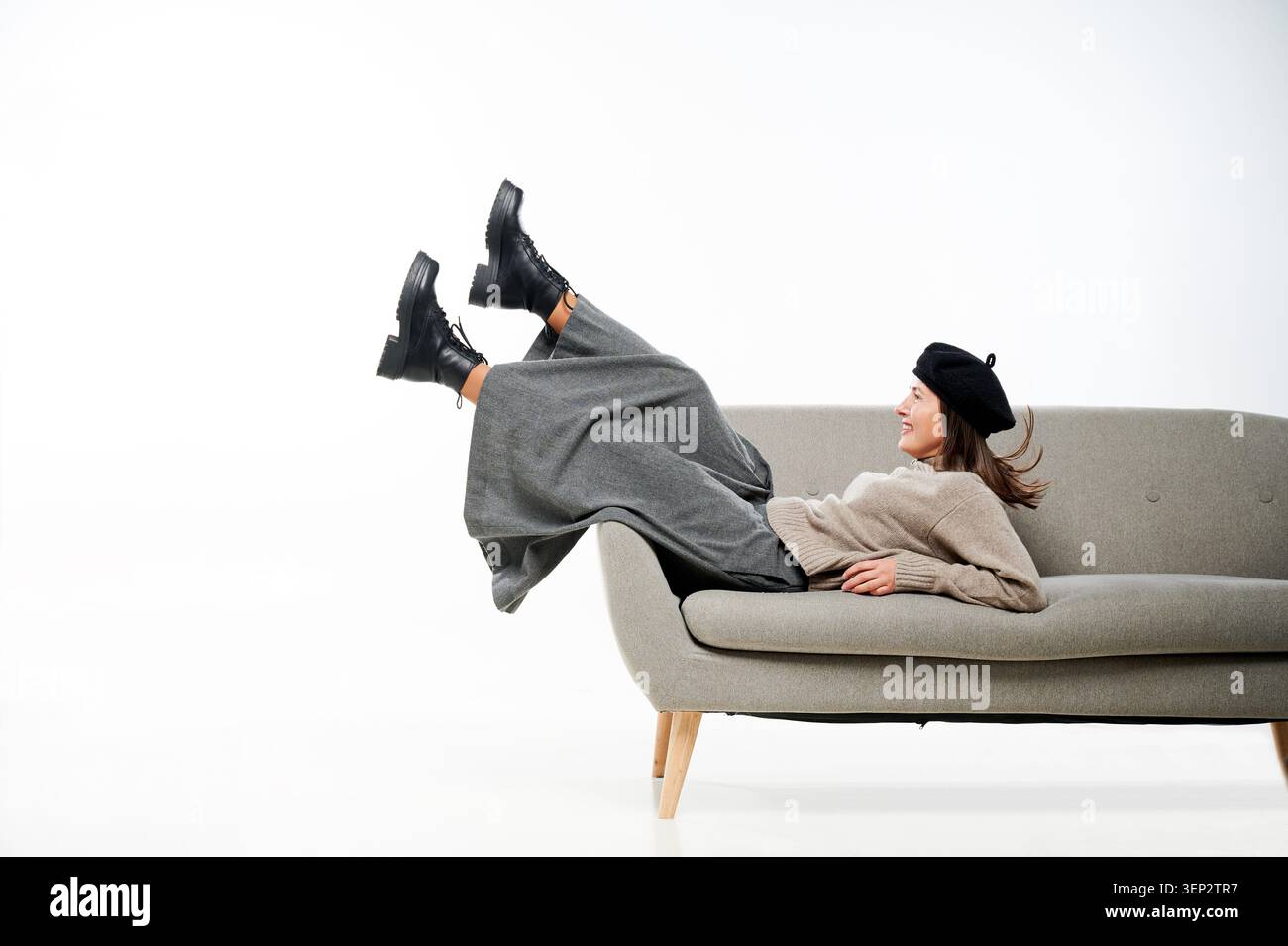 Donna sdraiata sul divano con gambe rialzate e sorridente in studio. Giovane donna caucasica che indossa berretto nero, maglione beige, pantaloni larghi grigi, stivali neri, umore rilassato e giocoso Foto Stock