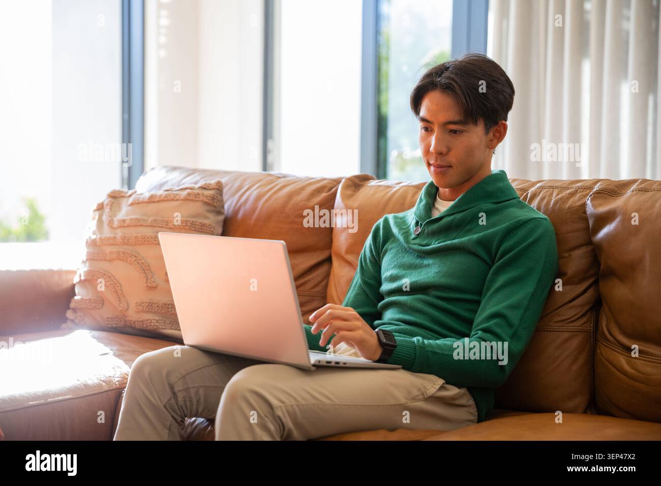 Uomo asiatico che indossa un pullover verde e pantaloni kaki, seduto su un divano abbronzante e lavora su un computer portatile d'argento Foto Stock