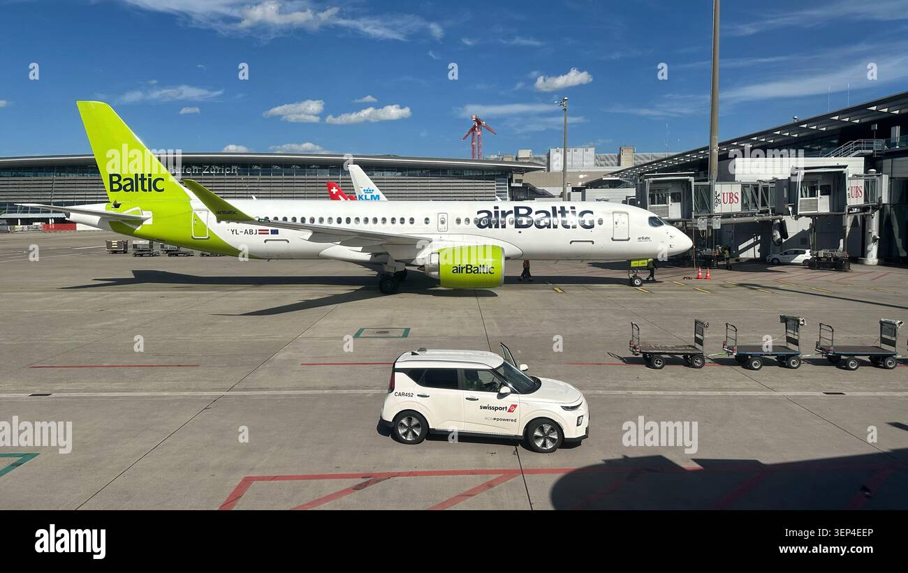 Zurigo, Svizzera - giugno 2025: AirBaltic Airbus A220-300 parcheggiato presso un cancello con i veicoli Swissport all'aeroporto di Zurigo. Foto Stock