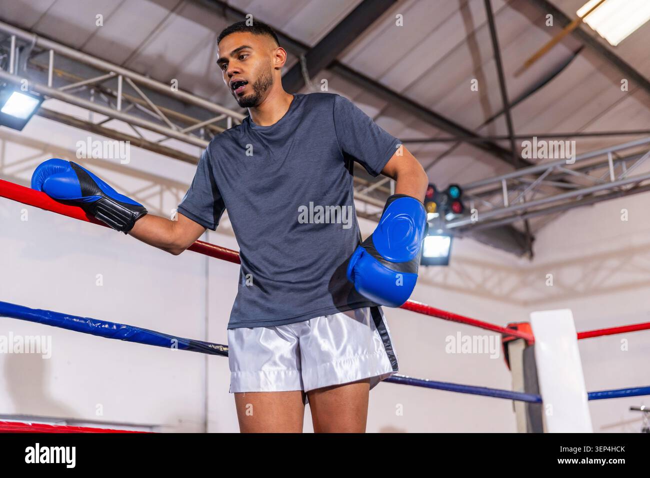 Uomo in pantaloncini bianchi che indossa guanti blu appoggiato su corde ad anello presso il palco dello sport sotto le luci del palcoscenico Foto Stock