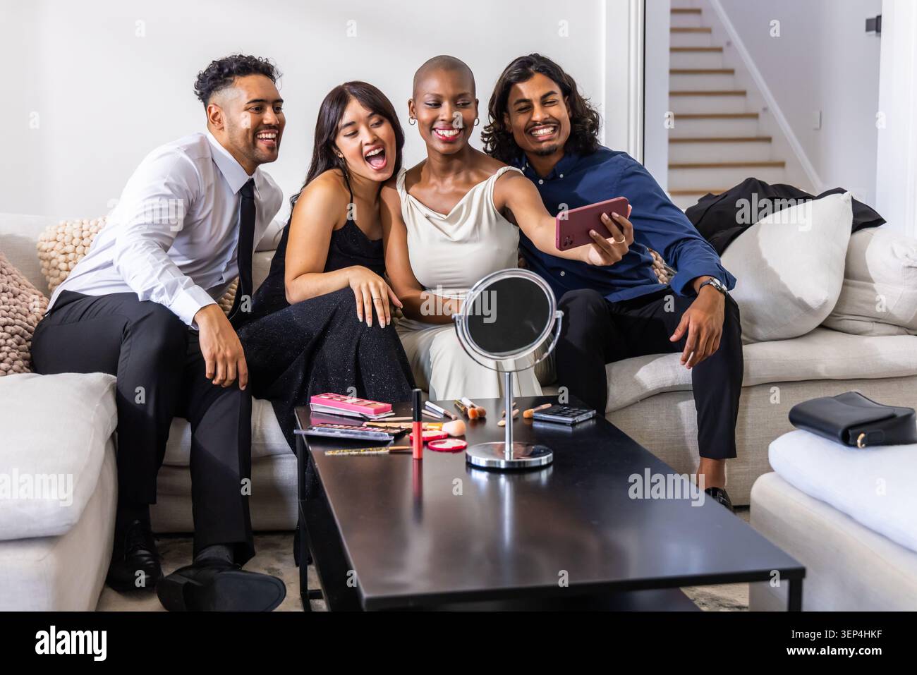 Amici diversi che posano per selfie sul divano, indossando abiti semi-formali con smartphone e trucco Foto Stock