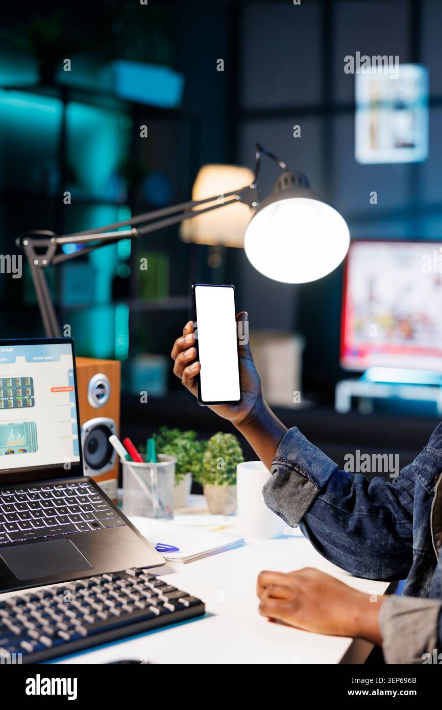 Donna afroamericana che lavora da casa su una scrivania con computer portatile e smartphone con schermo bianco, giovane che afferra verticalmente un dispositivo mobile che visualizza un modello cromakey isolato. Foto Stock