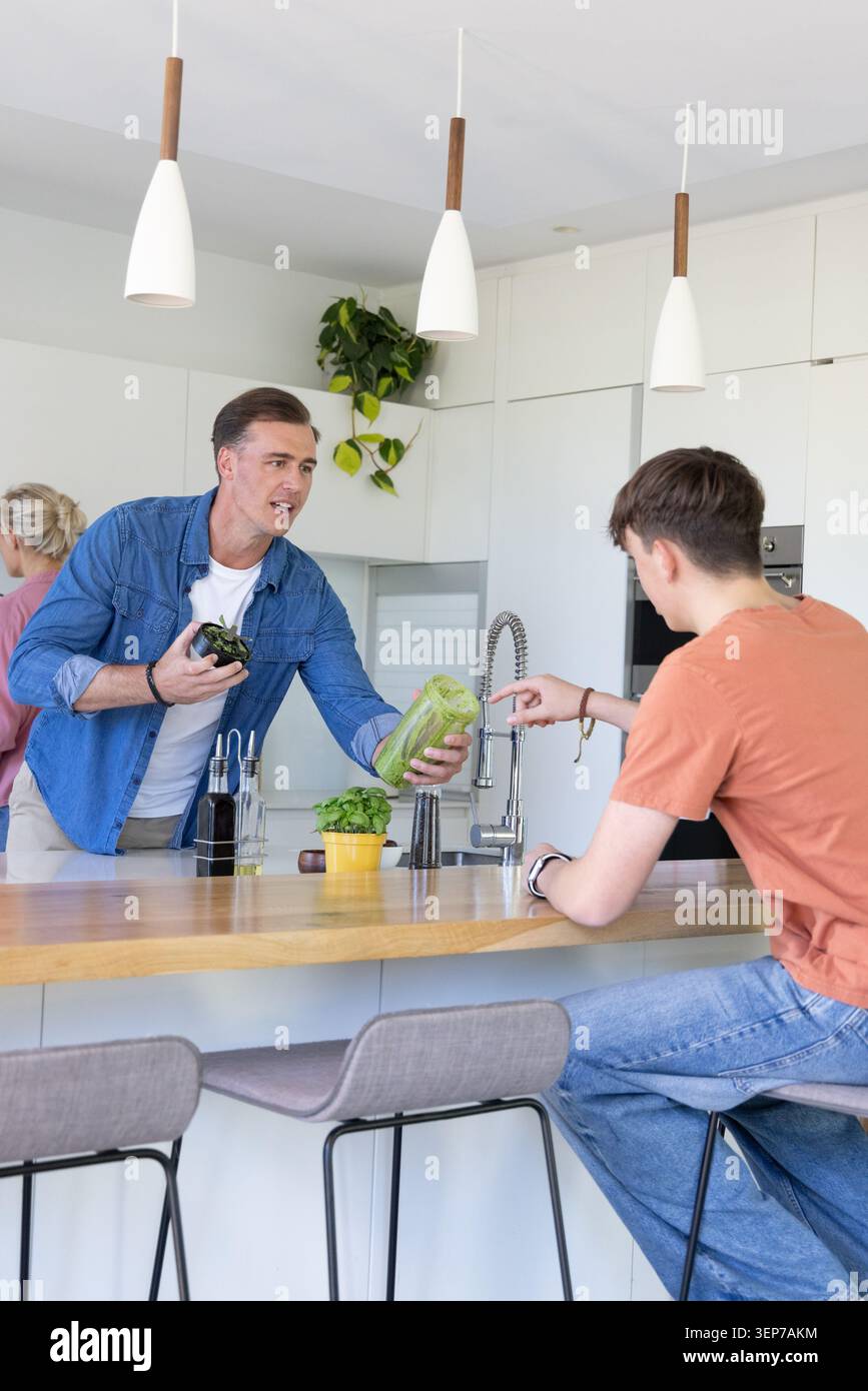Famiglia in cucina con camicia in denim che consegna lattuga ai giovani con t-shirt arancione al rubinetto Foto Stock