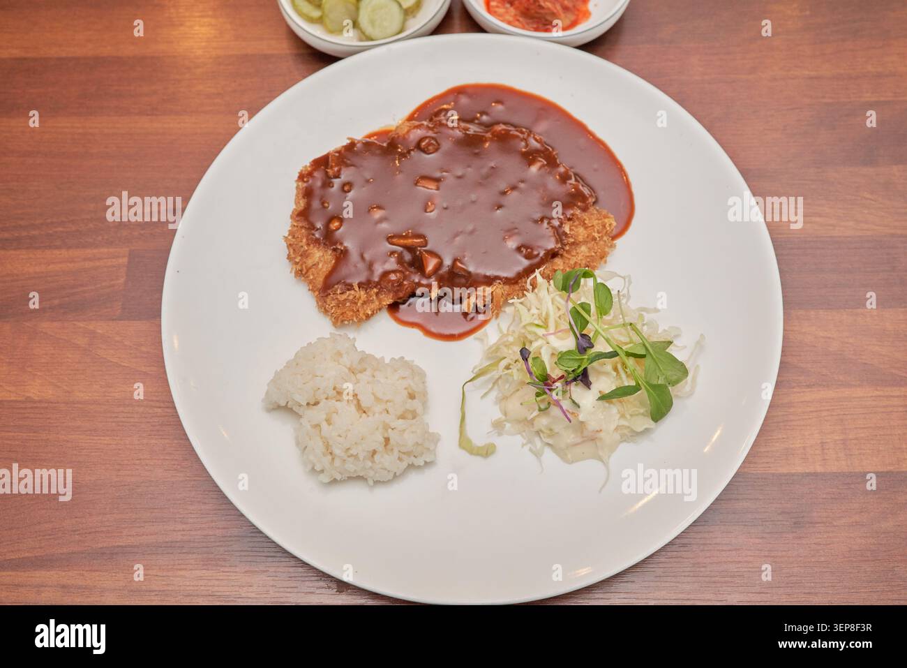 Vista dall'alto della cotoletta di maiale fritta ricoperta di salsa marrone scuro su un piatto bianco con riso al vapore e insalata di cavolo su un tavolo di legno Foto Stock