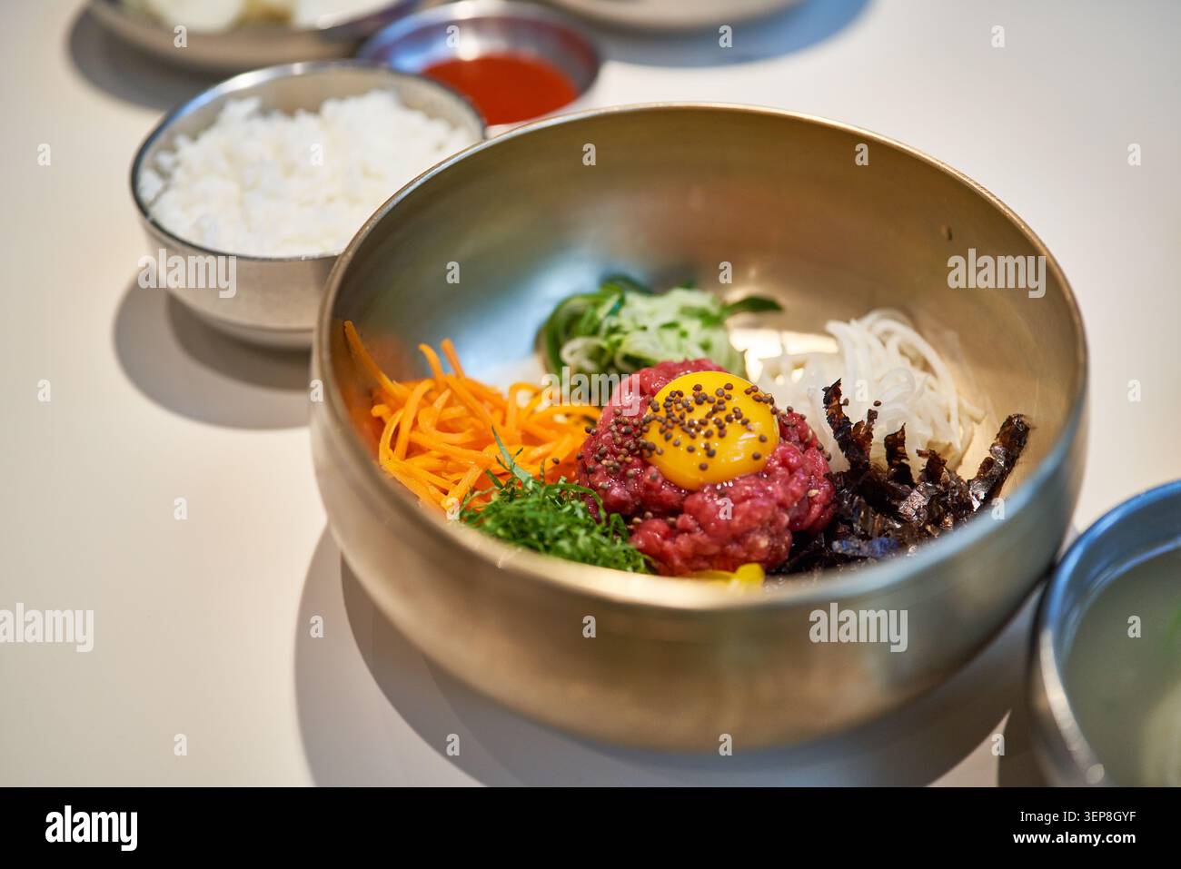 Bibimbap di manzo crudo asiatico tradizionale di prima qualità condito con tuorlo d'uovo e verdure fresche in una ciotola di ottone che mostra un'autentica cucina gourmet di alto livello. Foto Stock