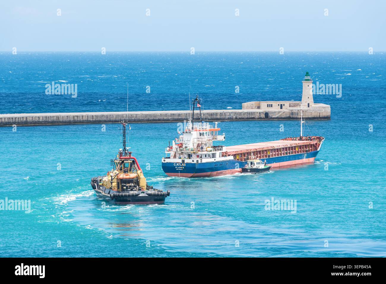 La Valletta, Malta - 16 maggio 2025: Nave da carico, rimorchiatore e pilota navigando vicino all'ingresso storico del porto e al faro di la Valletta, Malta, surro Foto Stock
