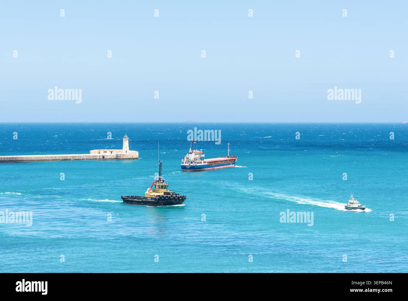 La Valletta, Malta - 16 maggio 2025: Nave da carico, rimorchiatore e pilota navigando vicino all'ingresso storico del porto e al faro di la Valletta, Malta, surro Foto Stock