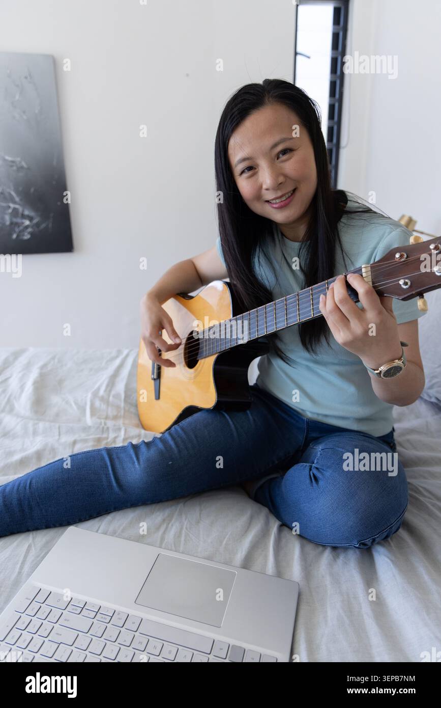 Donna asiatica seduta a gambe incrociate sul letto, che suona una chitarra acustica in legno chiaro con un laptop argentato Foto Stock
