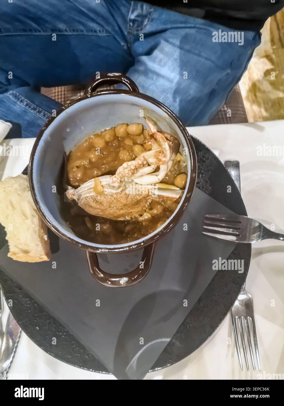 Primo piano di un'abbondante zuppa di granchi e fagioli servita in una pentola vintage. Vista dall'alto di un delizioso pasto a base di pesce con una persona seduta al tavolo - Immagine stock catturata con smartphone