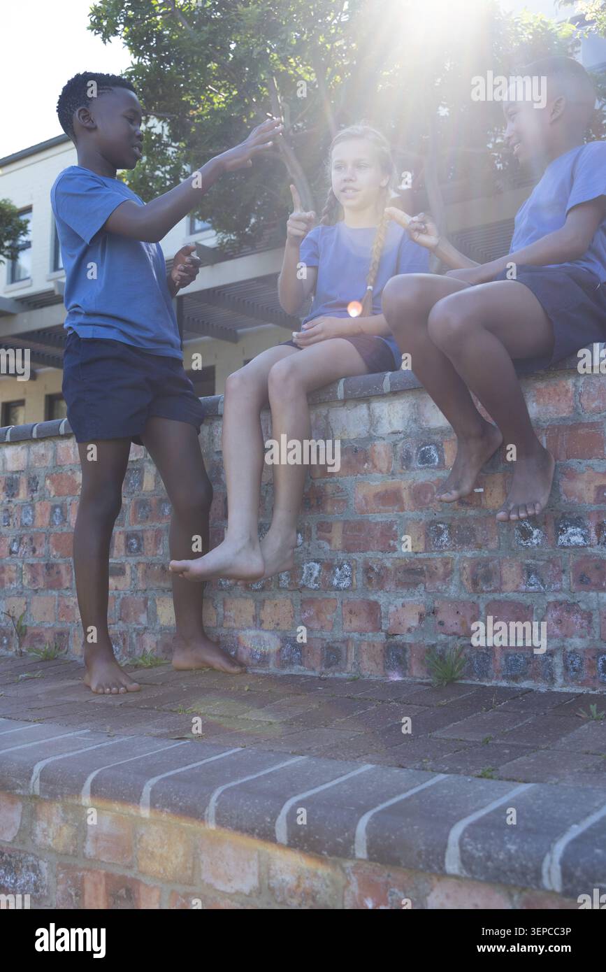 Tre bambini in età scolare che indossano magliette blu seduti su un muro di mattoni nel cortile della scuola, tenendo uno spuntino Foto Stock