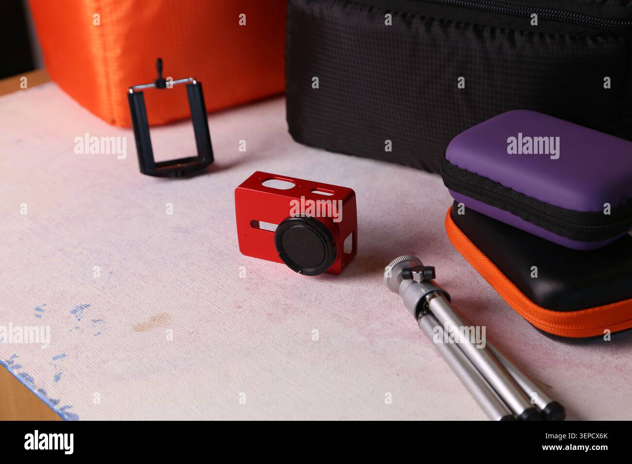 Un accessorio per fotocamera rosso si trova accanto alle custodie viola e nere su un tavolo. Nelle vicinanze, un piccolo treppiede e una clip sono in attesa di essere utilizzati. La scena mostra una configurazione per scattare foto. Foto Stock
