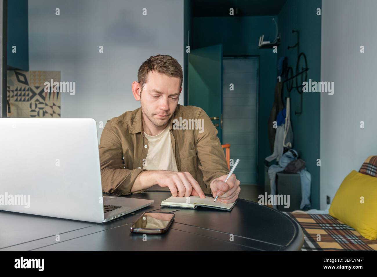 Uomo che scrive appunti su un notebook di carta mentre lavora su un computer portatile a casa. Nomade digitale maschile su lavoro remoto, pianificazione e produttività a casa Foto Stock