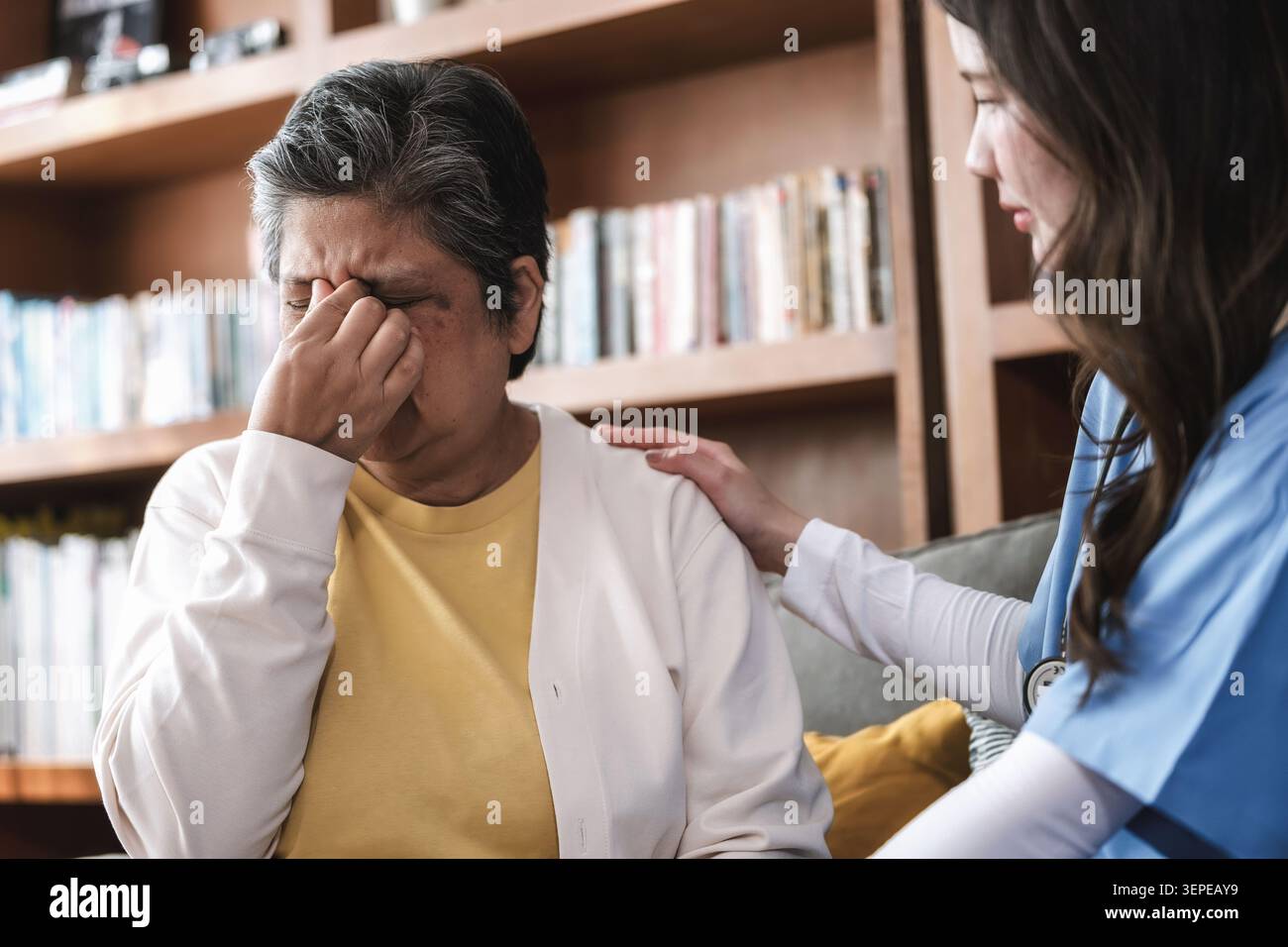 Una donna anziana strofina i templi nel dolore. Le sue spalle sono tese con disagio. La giovane badante le mette una mano confortante sulla spalla. Libreria linea Foto Stock