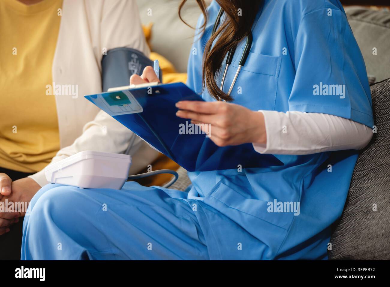 Infermiere in scrub blu controlla la pressione sanguigna, l'operatore sanitario tiene gli appunti mentre misura la pressione sanguigna con il bracciale sul paziente. Il paziente si siede comodamente Foto Stock