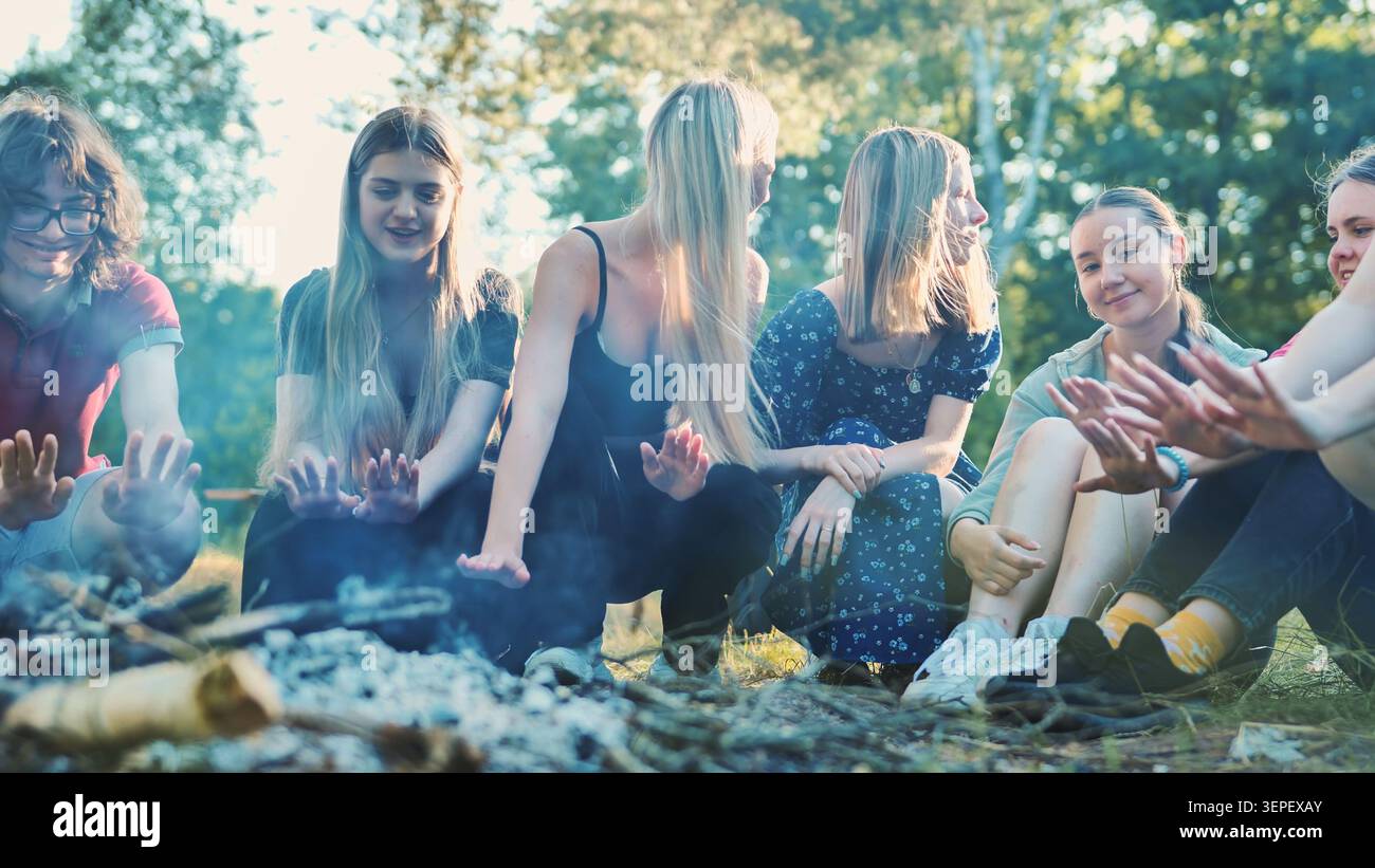 I giovani studenti si scaldano le mani vicino al falò scoppiettante, condividendo il cameratismo in mezzo al bosco Foto Stock
