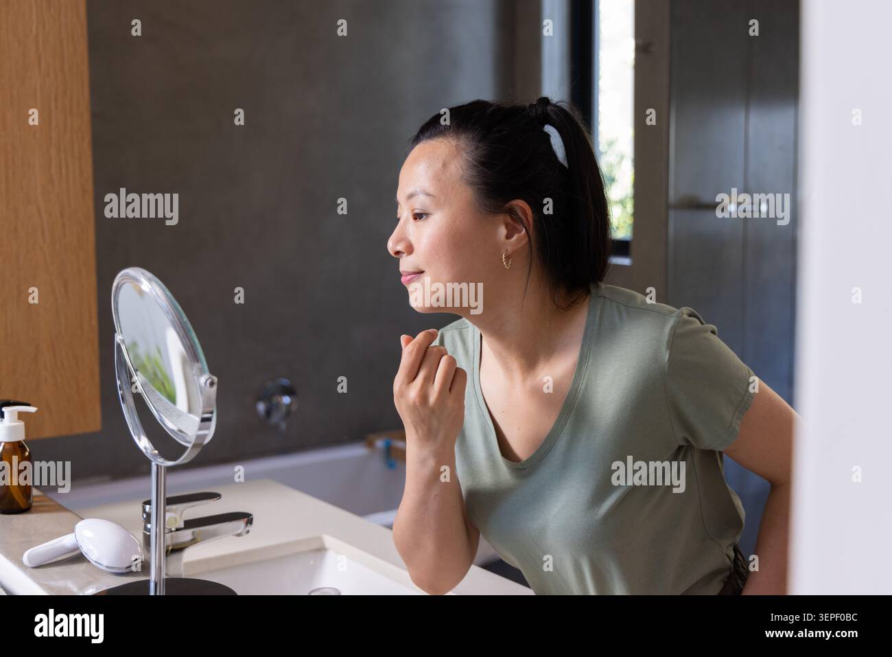 Donna asiatica appoggiata al lavandino del bagno, toccando il viso nello specchio rotondo con dispositivo per il viso nelle vicinanze Foto Stock