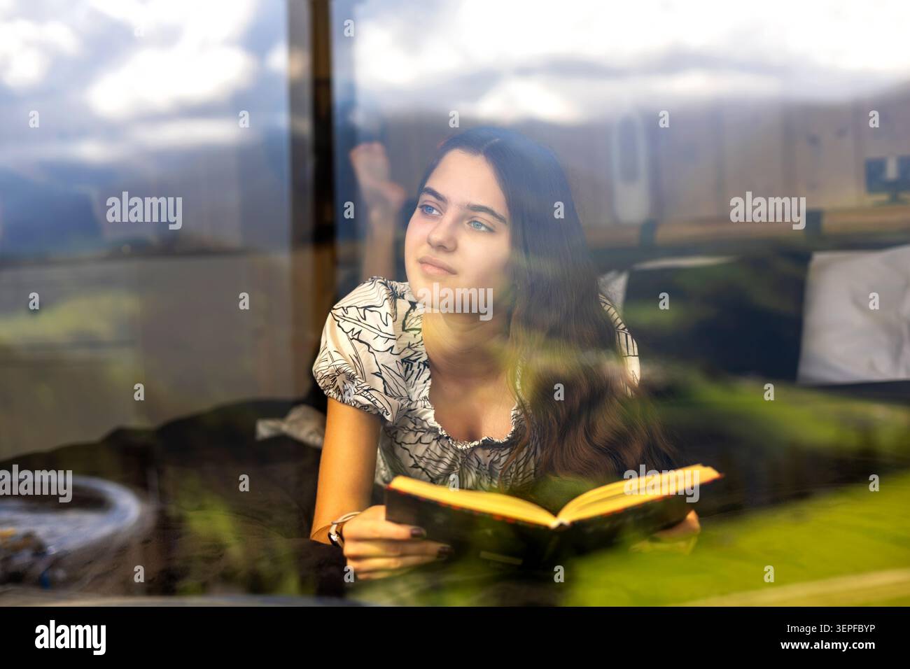 Giovane donna premurosa che legge da una finestra riflettente. Giovane donna sdraiata accanto a una finestra con un libro aperto, che guarda con attenzione attraverso il vetro riflettente. Foto Stock