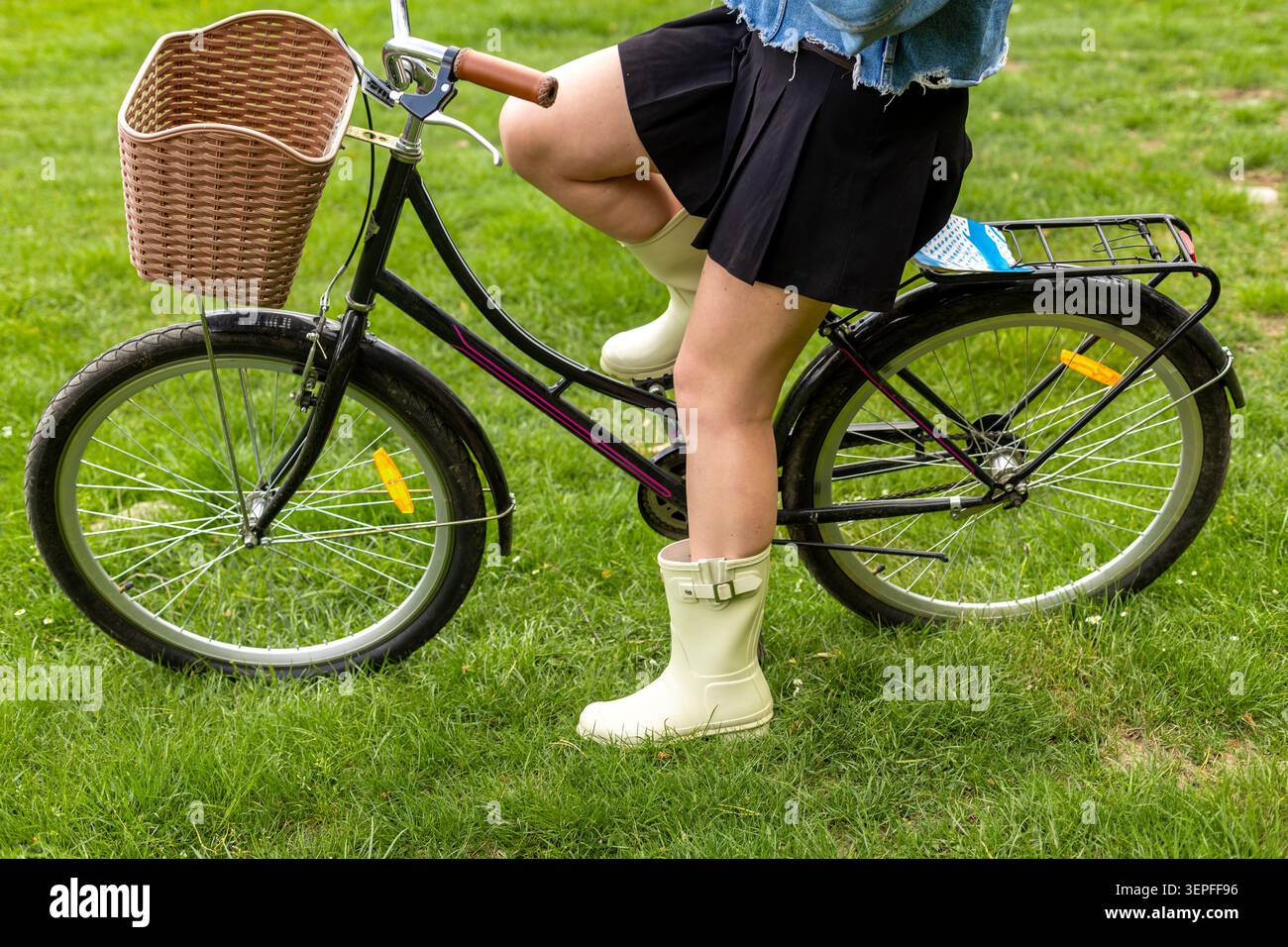 Giovane donna in bicicletta con cesto di vimini nel parco. Primo piano di una giovane donna seduta su una bicicletta nera con un cesto anteriore in vimini su erba verde. Foto Stock