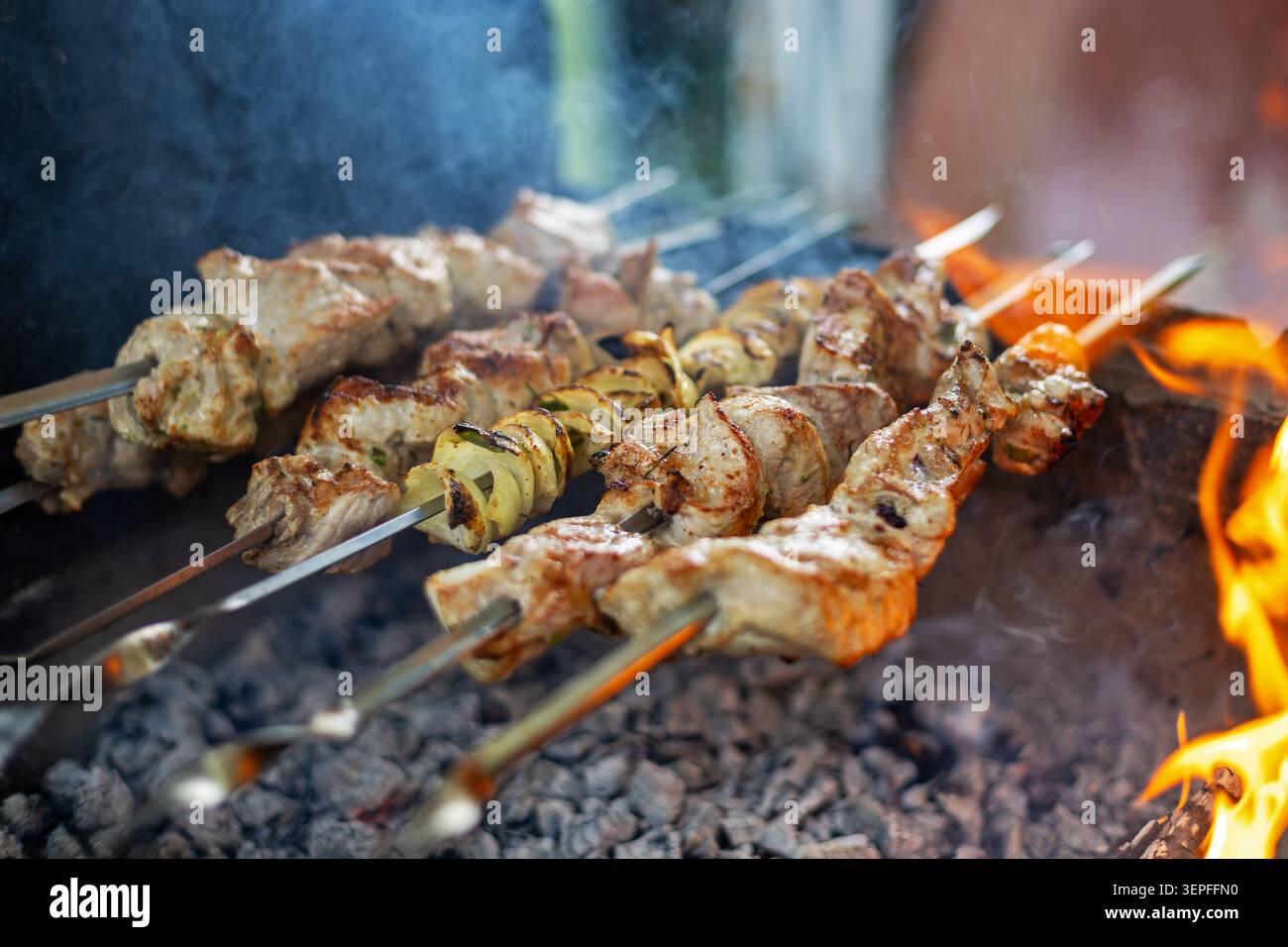 Spiedini di carne grigliati con cottura a fuoco aperto. Spiedini di metallo con carne alla griglia e verdure a fette cucinano su carbone caldo accanto alle fiamme brillanti. Foto Stock