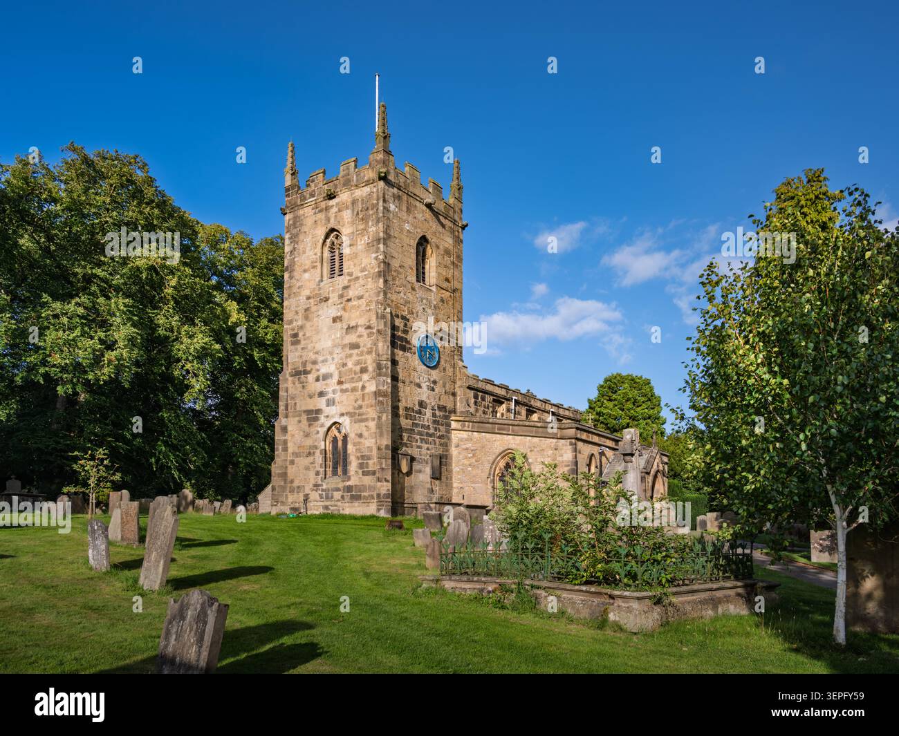 Splendida vista del sole che mette in risalto la maestosa torre in pietra e i giardini curati della chiesa parrocchiale di Eyam sotto un cielo azzurro Foto Stock