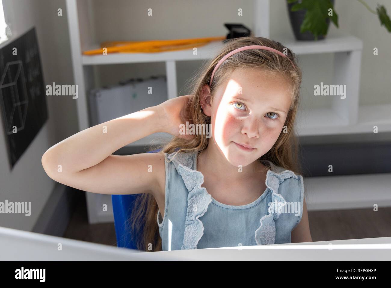 Ragazza preadolescente seduta alla scrivania bianca che tocca il collo nello studio con un toppa alla luce del sole e un abito azzurro Foto Stock