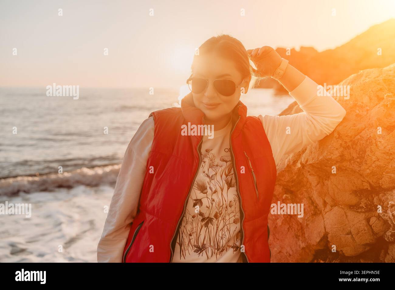 Donna spiaggia tramonto elegante signora che posa tra le rocce costiere indossando un gilet rosso e aviatori durante una calda ora dorata Foto Stock