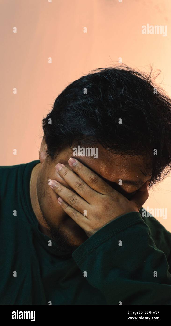 Un uomo adulto in difficoltà che indossa una camicia verde scuro inchina la testa e copre gli occhi con una mano per mostrare sentimenti di tristezza o di profonda stanchezza Foto Stock