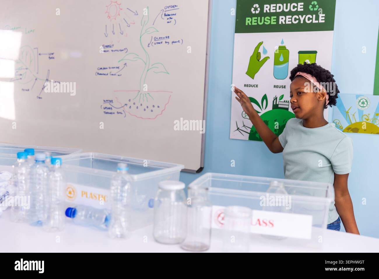 Ragazza afroamericana che indica i contenitori di riciclaggio in classe con bottiglie e lavagna bianca, spazio copia Foto Stock
