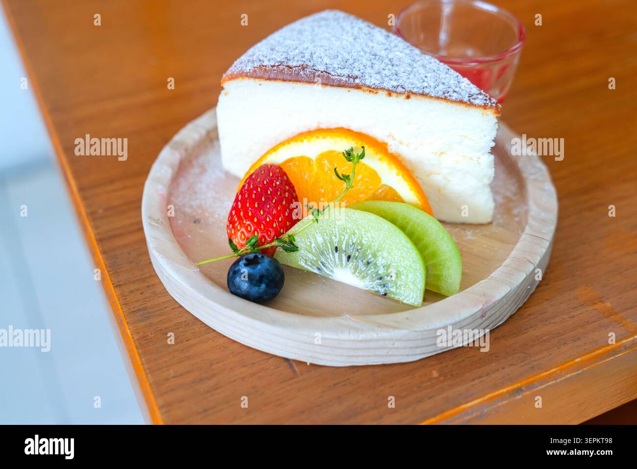Una fetta di cheesecake cremosa ricoperta di zucchero a velo e accompagnata da frutta fresca, tra cui fragole, mirtilli, kiwi e arancia Foto Stock