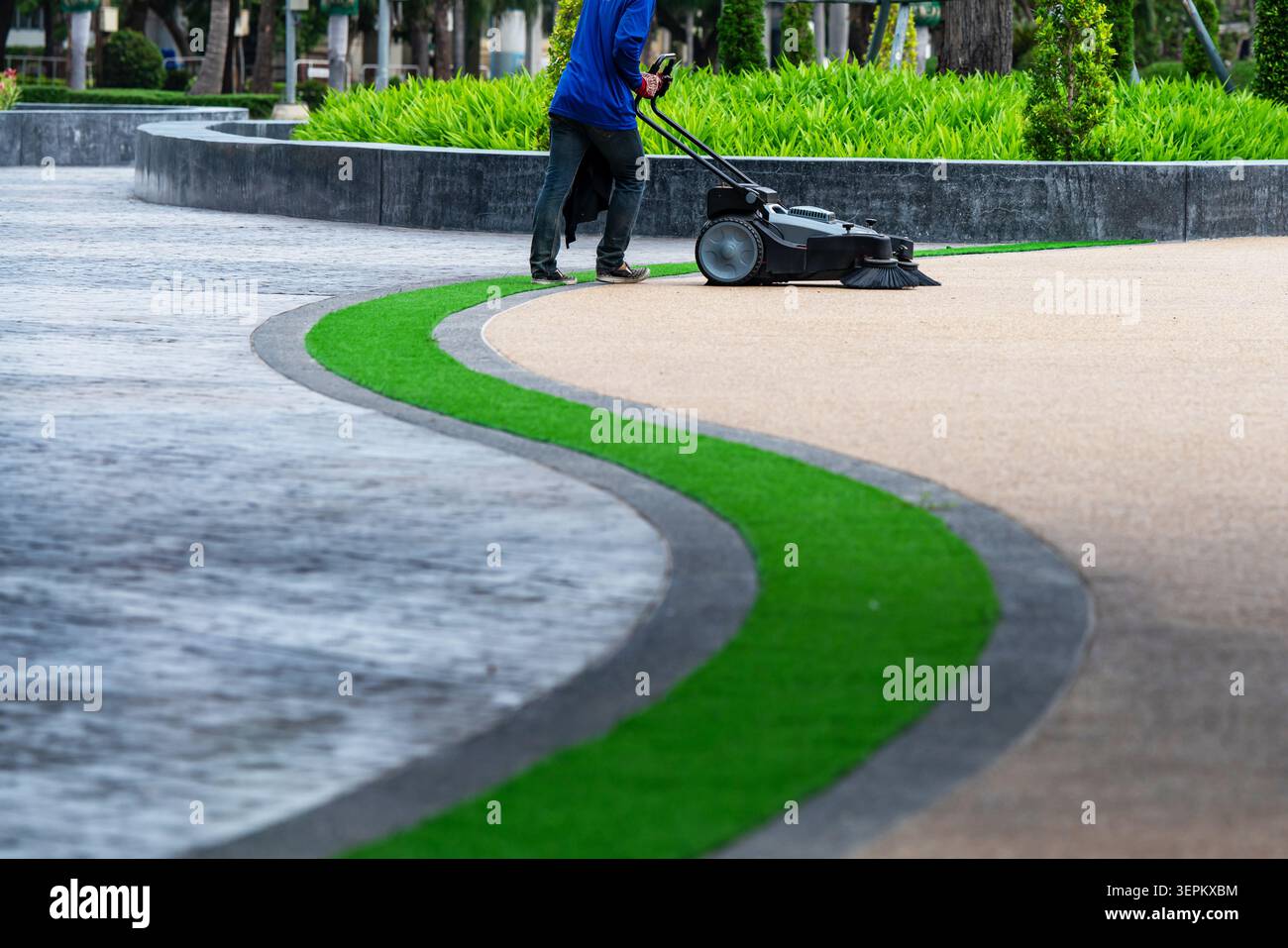 Un lavoratore sta gestendo un rasaerba lungo un percorso tortuoso in un parco lussureggiante, mantenendo il paesaggio verde con attrezzature di sicurezza e attrezzature professionali. Foto Stock