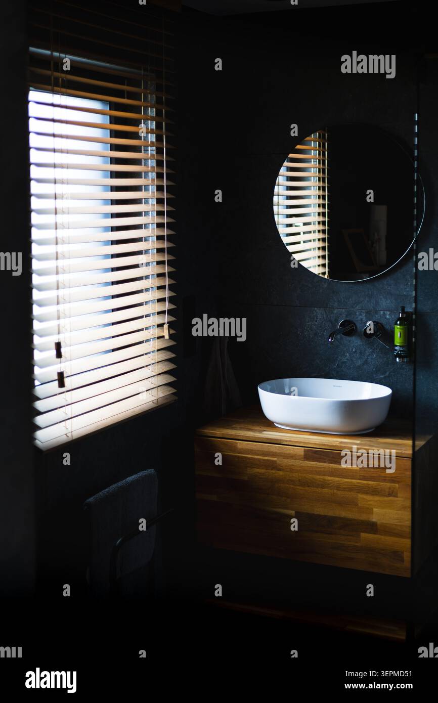 Gli interni del bagno sono moderni e minimalisti e presentano un lavandino in ceramica bianca su un'unità mobile in legno galleggiante, uno specchio a parete rotonda e naturale Foto Stock