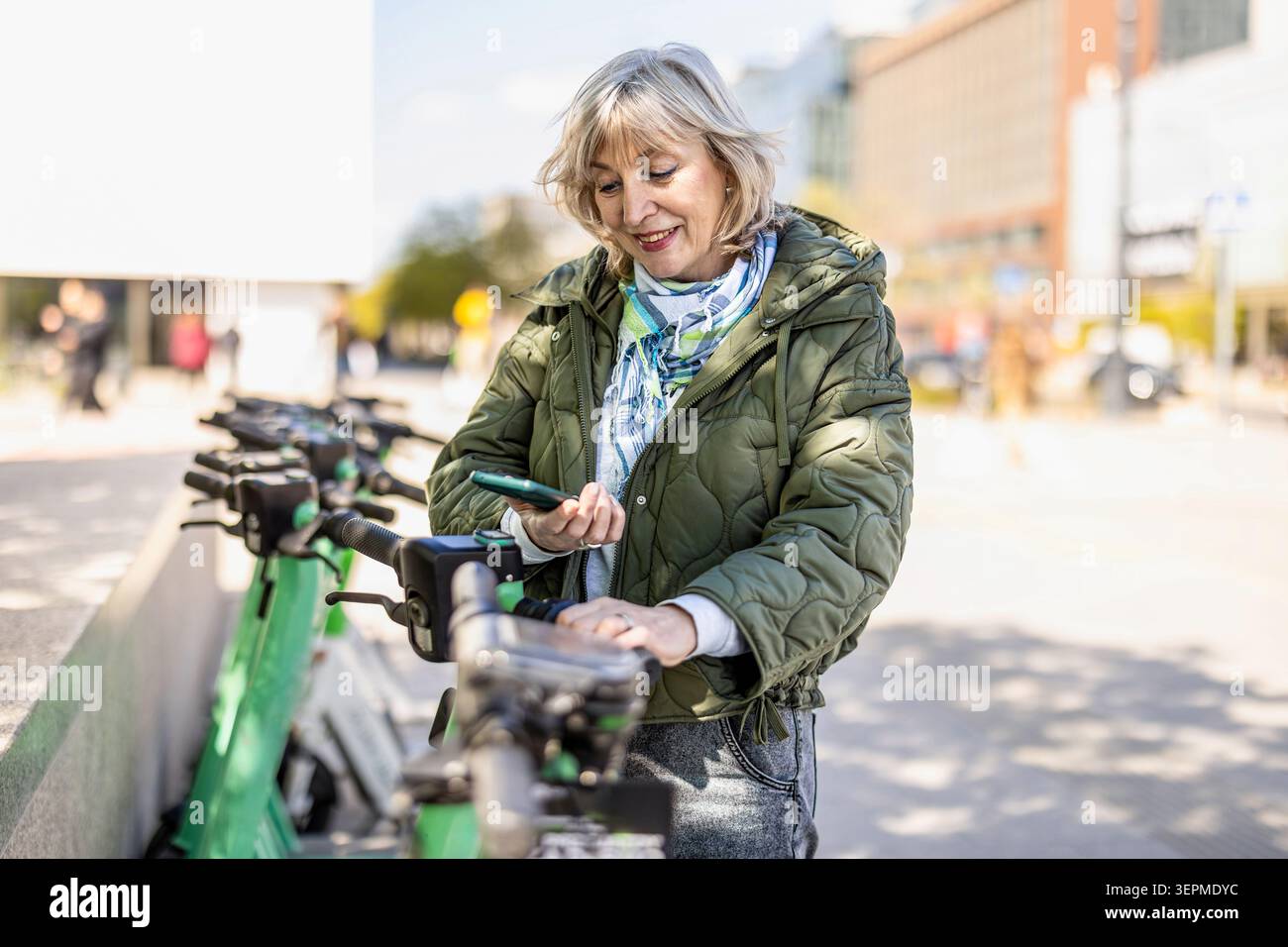 Donna matura che usa un'app per telefono per noleggiare uno scooter elettrico da città Foto Stock