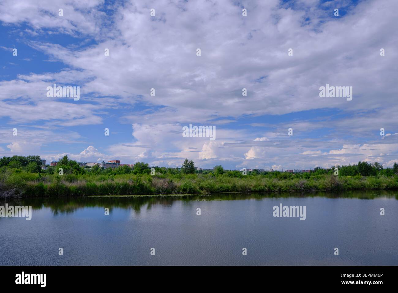 Lago di superficie dall'acqua calma che riflette nuvole luminose con alberi verdi ed edifici lontani Foto Stock