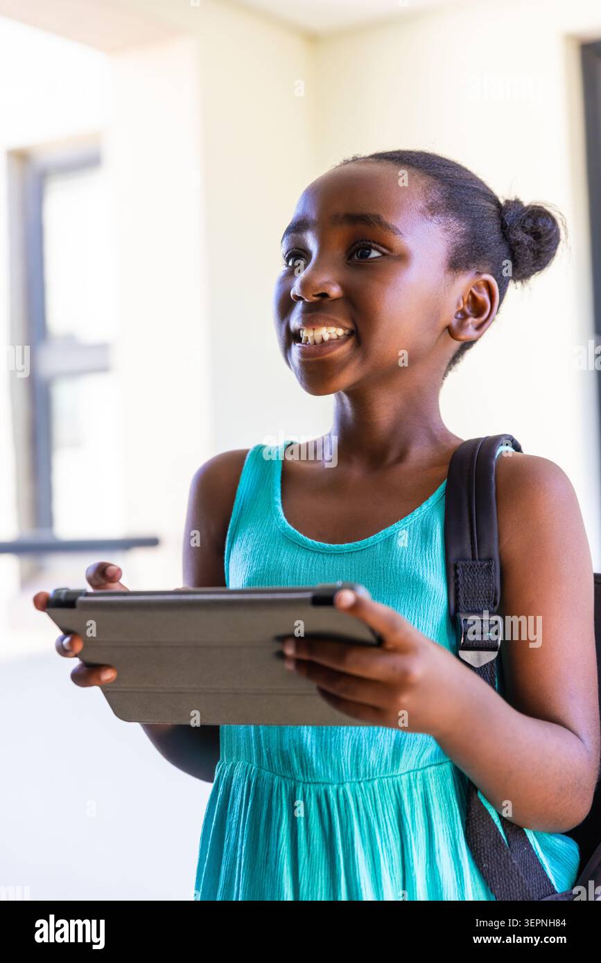 Bambino afroamericano sorridente e con un tablet in folio, che indossa un abito senza maniche di color verde acqua a scuola Foto Stock