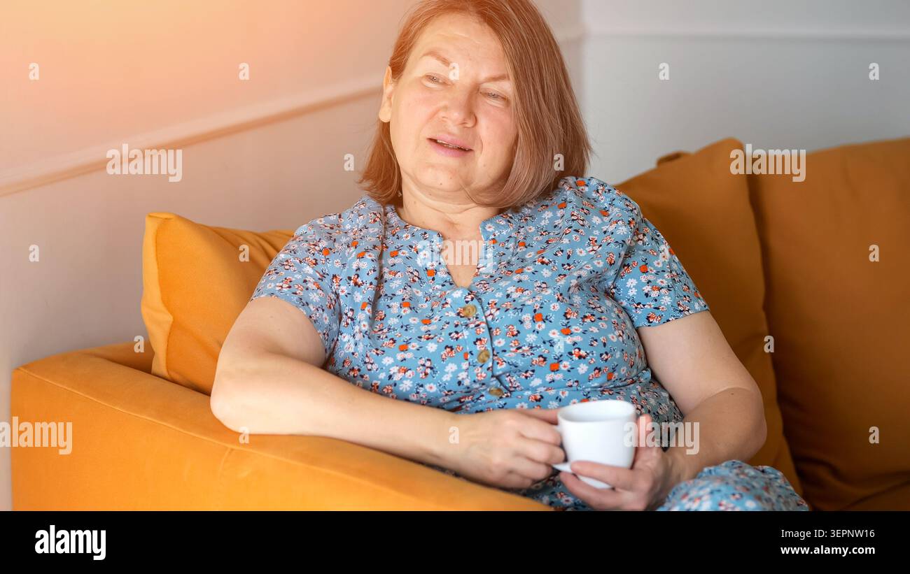 Serene Lady che si gode un pomeriggio accogliente. Donna rilassata seduta con calma e luce solare calda. Scena casual di donna che riposa comodamente sul divano arancione con Foto Stock