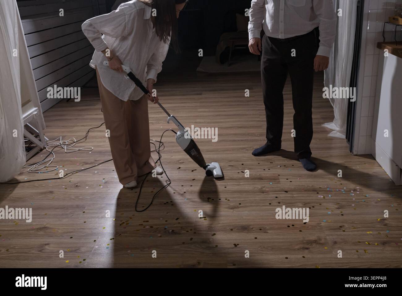 Aspirazione del pavimento in legno dopo il compleanno felice, persona in camicia bianca e pantaloni scuri che puliscono le briciole vicino al bordo del tavolo, movimento focalizzato degli strumenti di pulizia Foto Stock