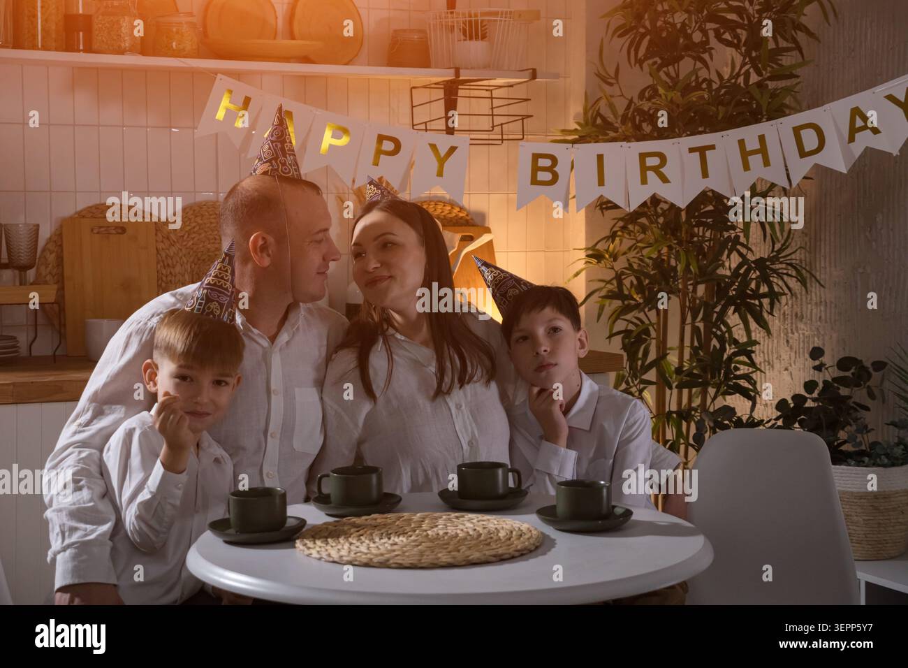 Festa di compleanno in famiglia al tavolo da cucina, calda scena serale con genitori e bambini che indossano cappelli da festa, banner di buon compleanno sullo sfondo Foto Stock