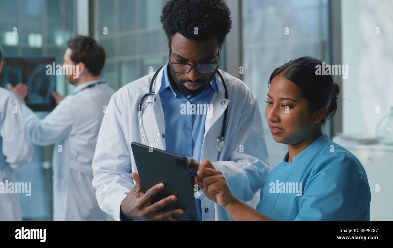 Due medici multirazziali che discutono la diagnosi guardando in tablet digitale in clinica. Uomo afroamericano medico con donna araba indiana infermiera che parla Foto Stock