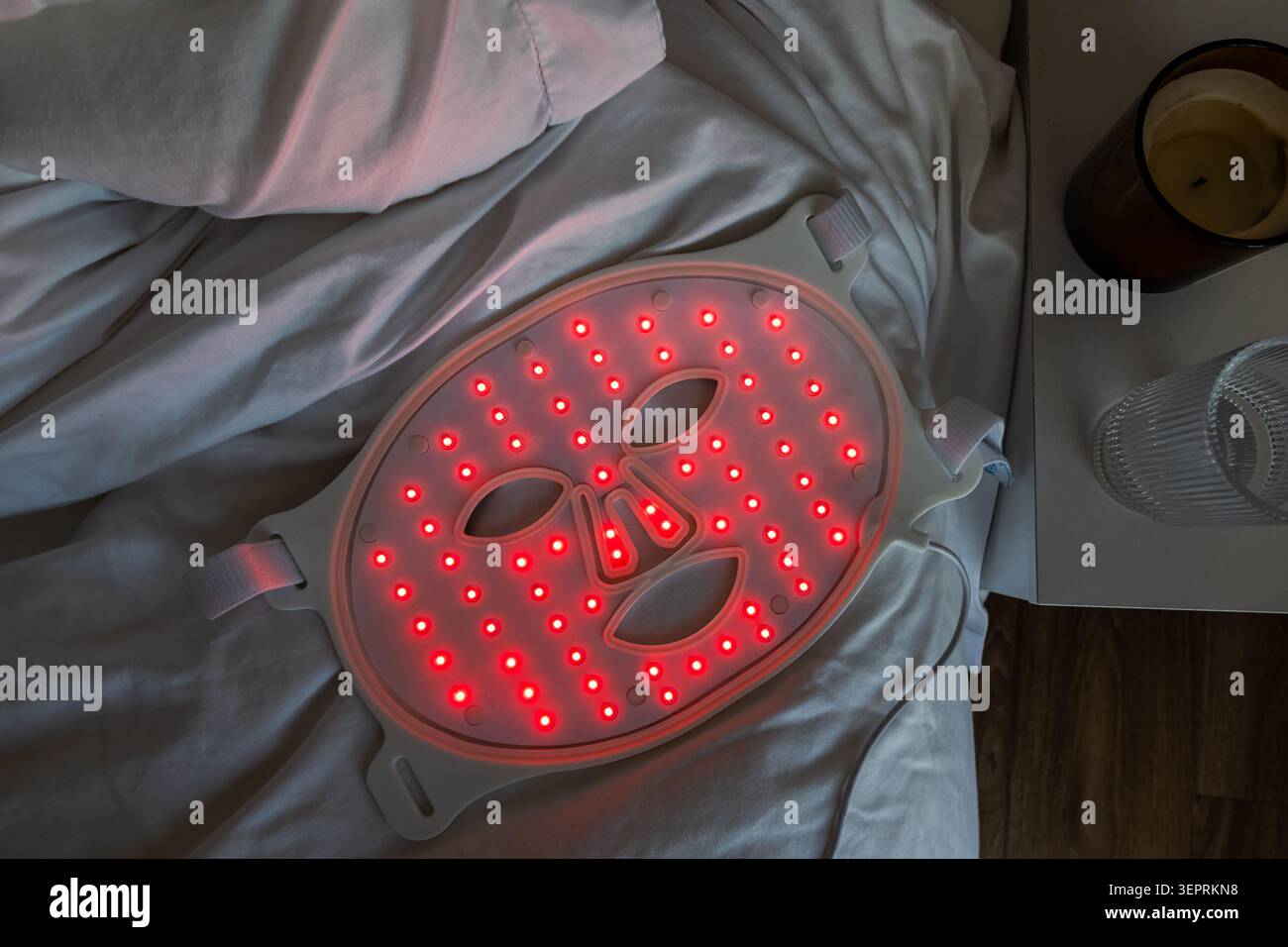Maschera per la terapia a luce rossa che poggia su lenzuola bianche accanto a una candela e un vetro, creando una tranquilla scena di cura della pelle e di auto con serate soffici Foto Stock