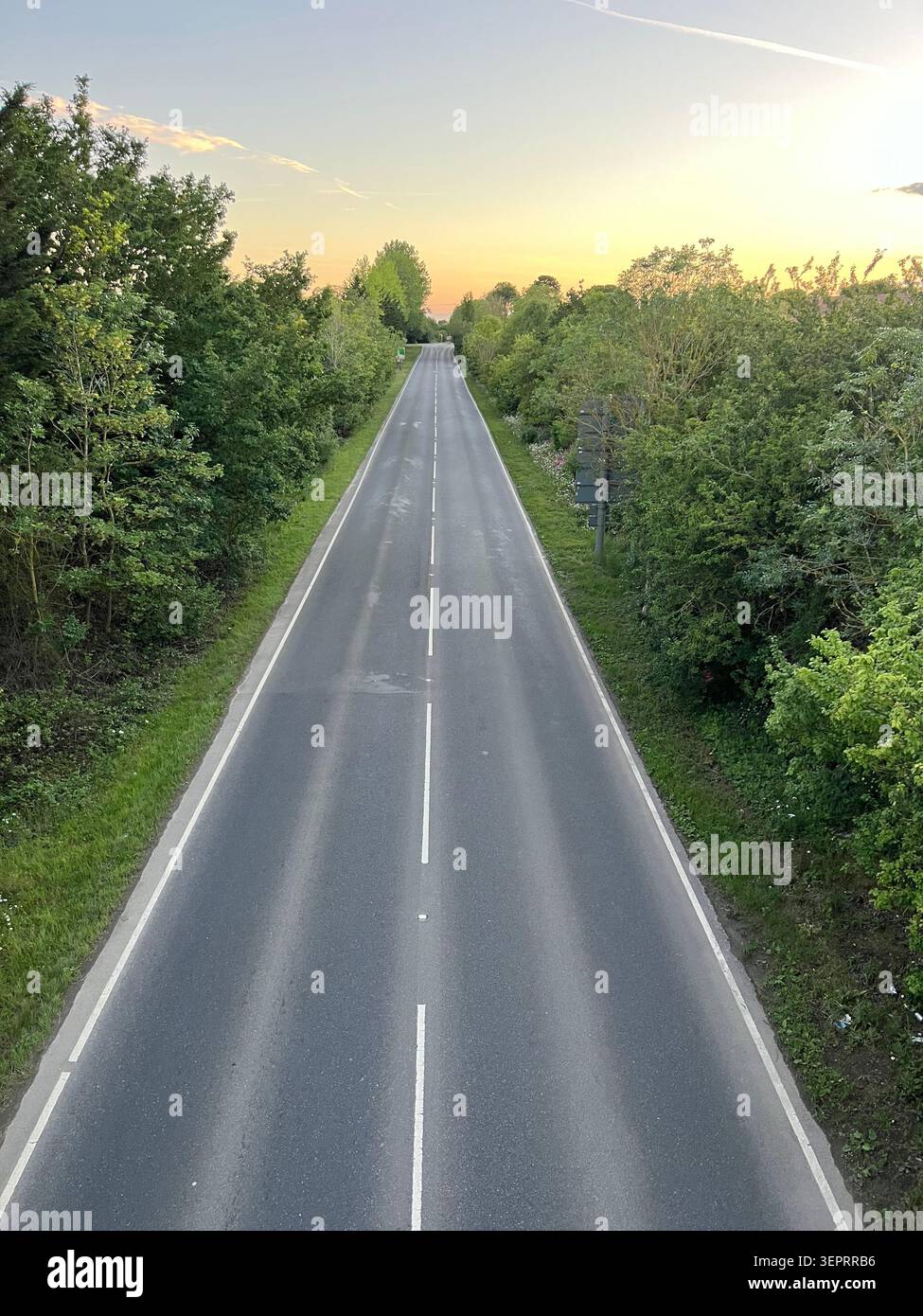 Un tratto tranquillo e vuoto del Downham Market bypass a Norfolk, visto da un ponte pedonale e circondato da alberi verdi sotto la luce soffusa della sera. - Immagine stock catturata con smartphone