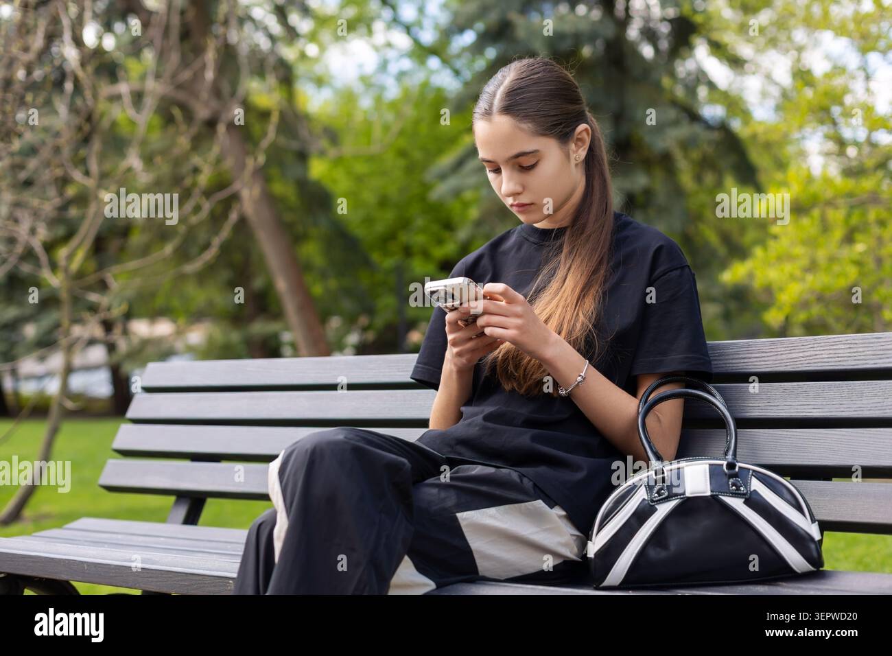 Giovane donna che usa lo smartphone sulla panca del parco. Una giovane donna siede su una panchina di legno del parco utilizzando uno smartphone, con una borsa nera accanto e un verde tenue Foto Stock