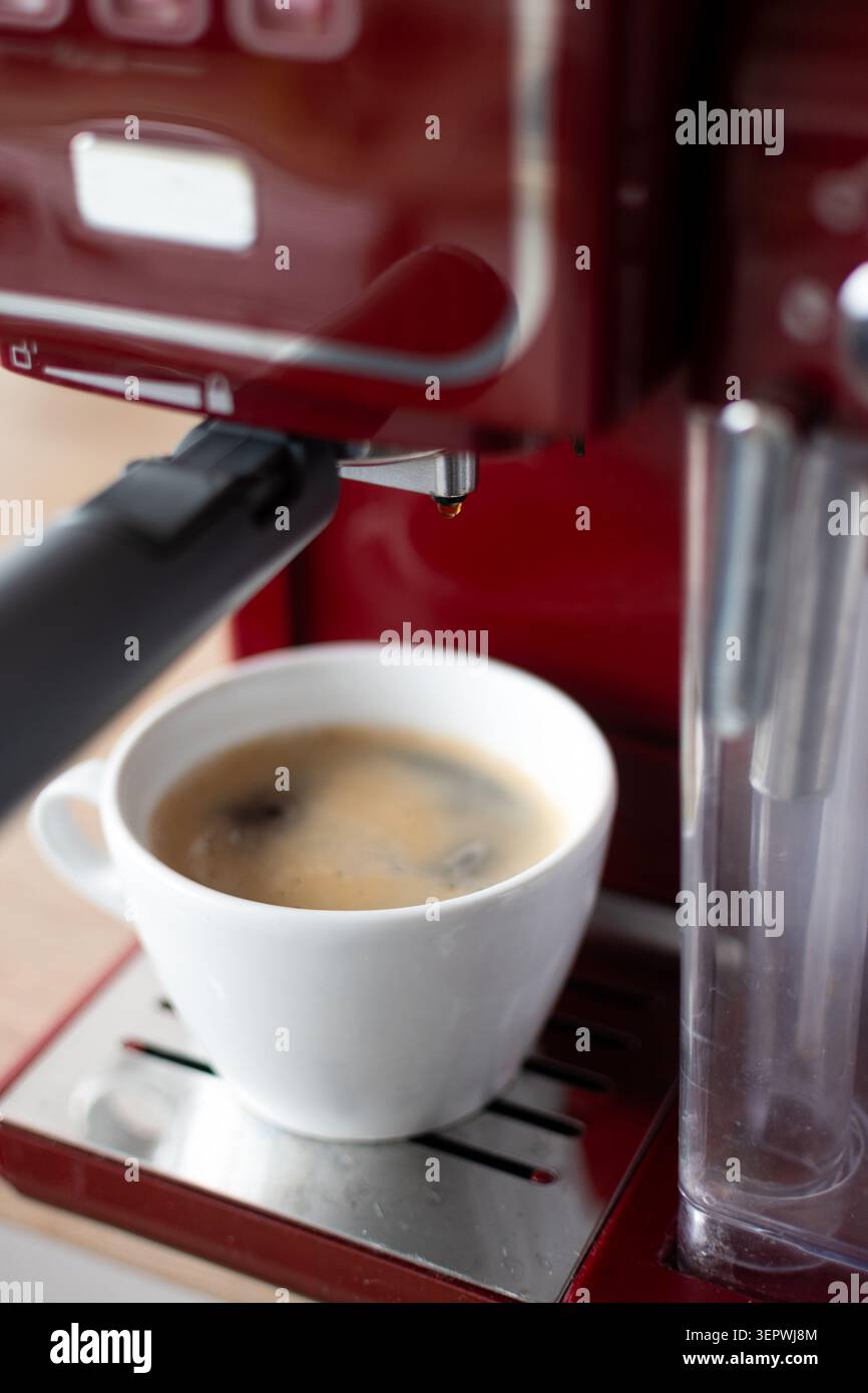 Primo piano di un espresso appena estratto servito in una tazza di ceramica bianca sotto una macchina da caffè rossa. Foto Stock