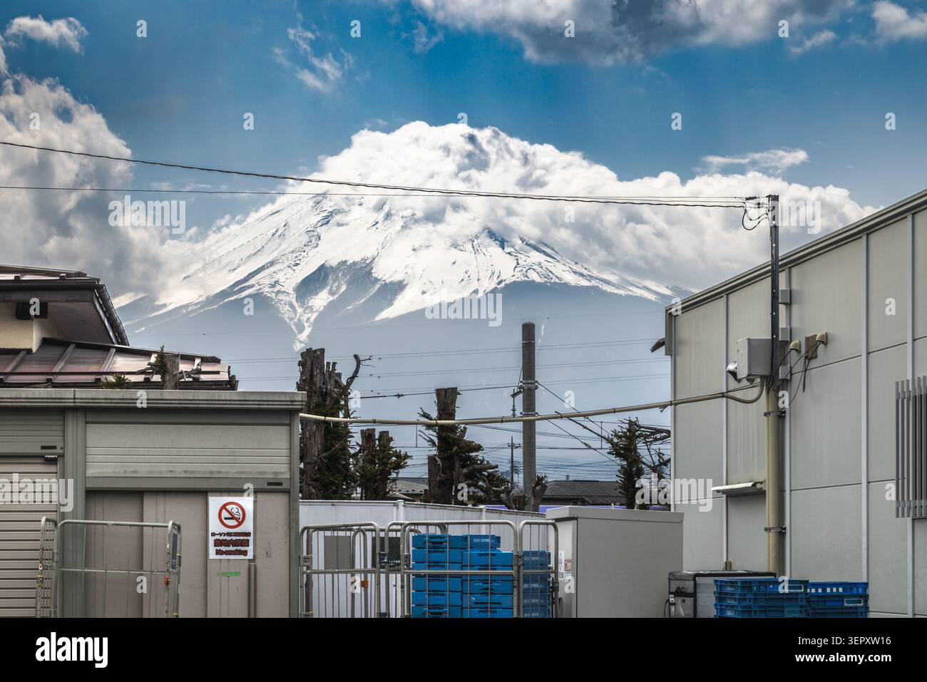 Il Monte Fuji avvolto dalle nuvole si erge oltre gli edifici industriali bassi e i cavi elettrici a Fujikawaguchiko, Yamanashi, Giappone. Foto Stock