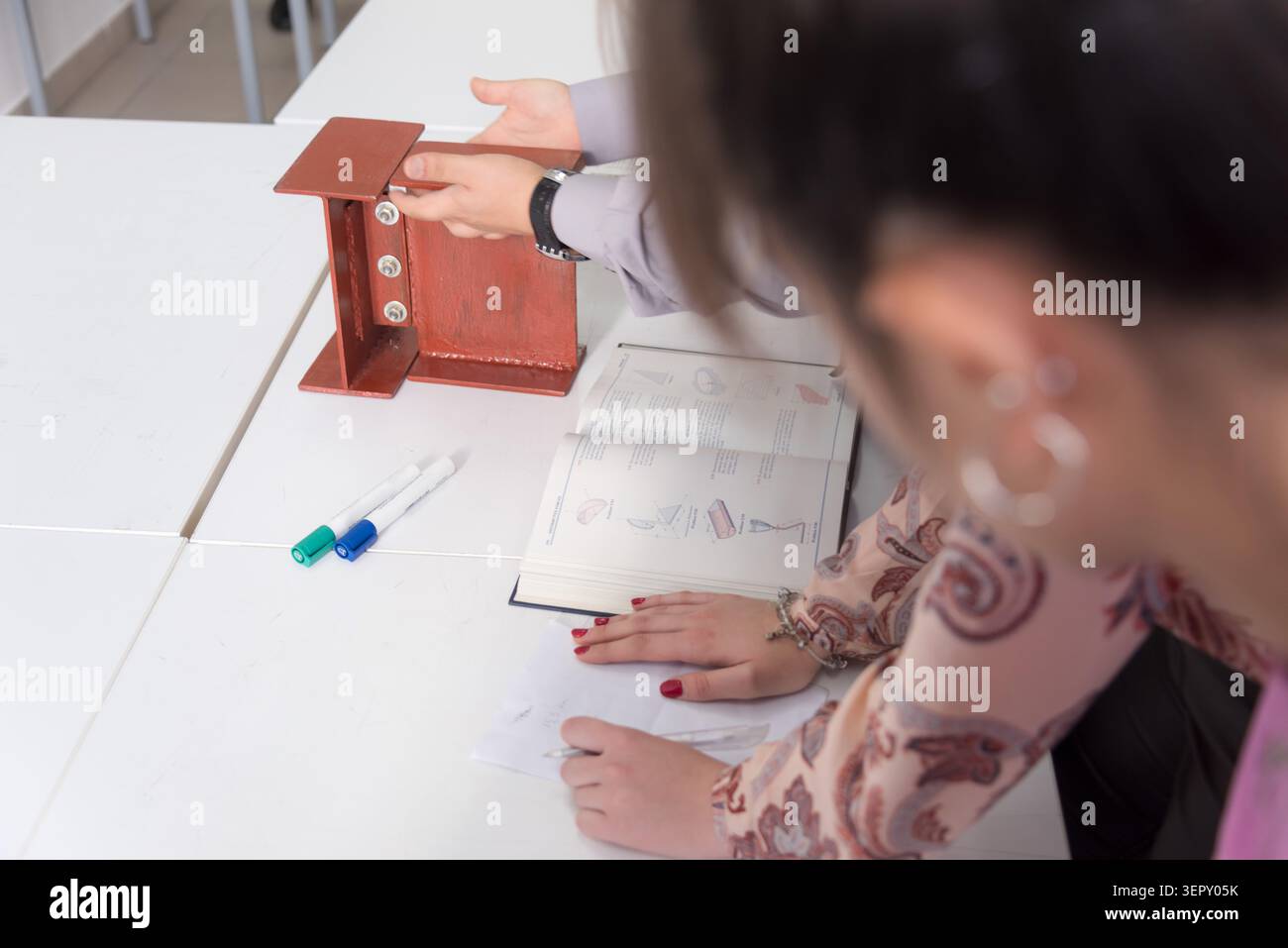 Uno scatto mirato che mostra le mani che manipolano due travi a i in acciaio rosso in miniatura su un tavolo bianco, utilizzato come ausilio visivo per l'insegnamento dell'ingegneria strutturale A. Foto Stock