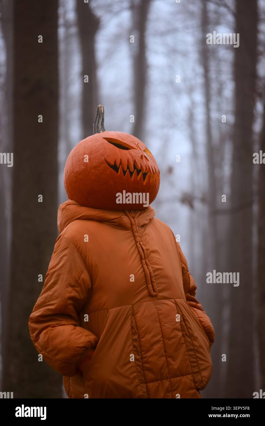 Primo piano ritratto di una figura misteriosa con una testa di Jack o lanterna che indossa una giacca arancione. Costume creativo di Halloween in un ambiente boscoso con atmosfera cinematografica Foto Stock