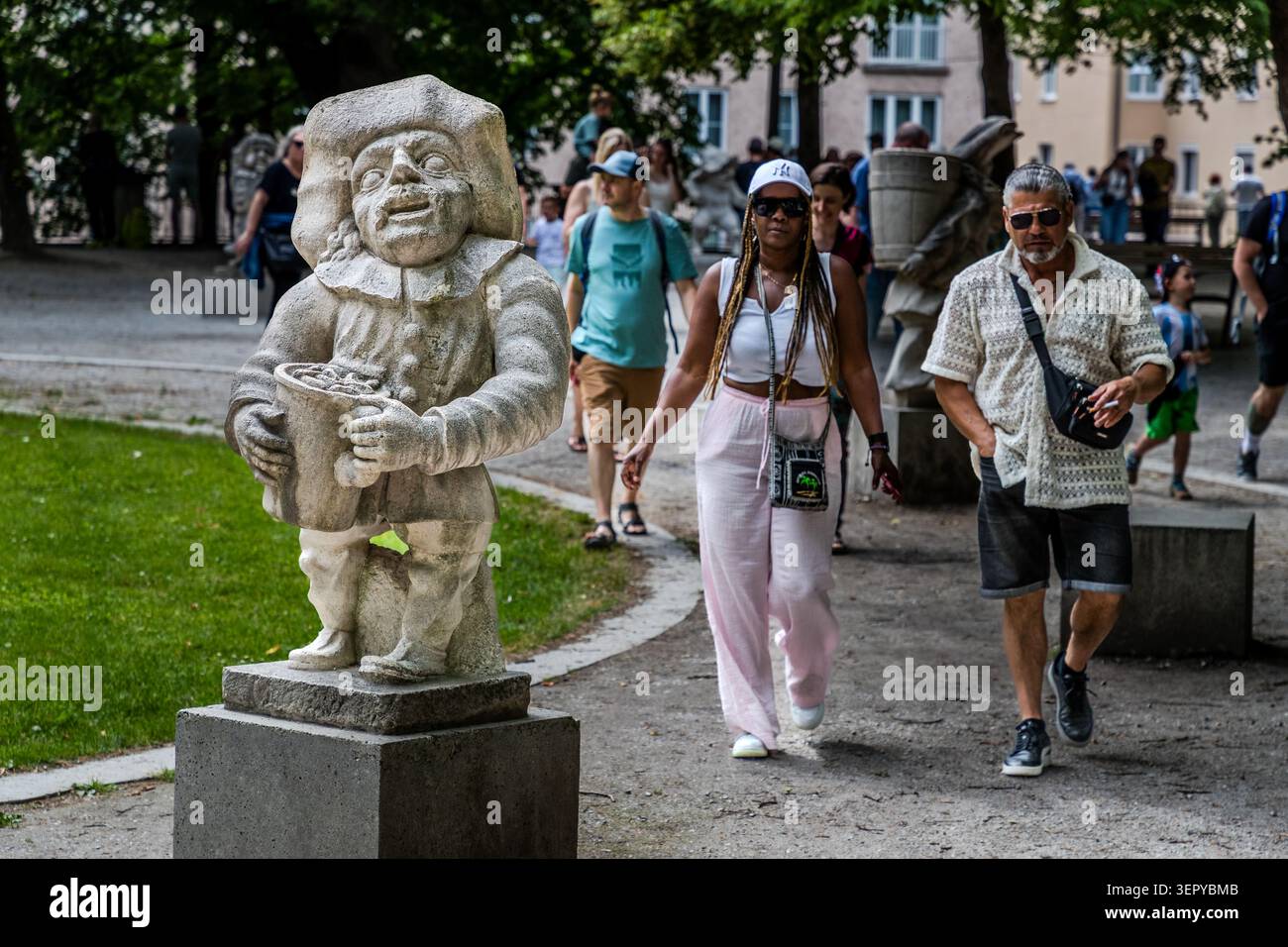 Una statua in pietra barocca di un nano che indossa un cappello a tegole larghe e porta un cesto di frutta nel Giardino Nano dei Giardini Mirabell a Salisburgo, con i turisti che passano in primo piano. La scena illustra l'elevato numero di visitatori dei giardini Mirabell, una delle attrazioni più famose di Salisburgo. Giardini Mirabell, Salisburgo, Austria Foto Stock